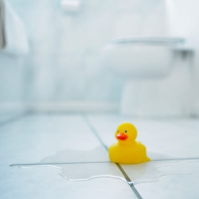 Small yellow rubber duck floating in a puddle on a tiled bathroom floor